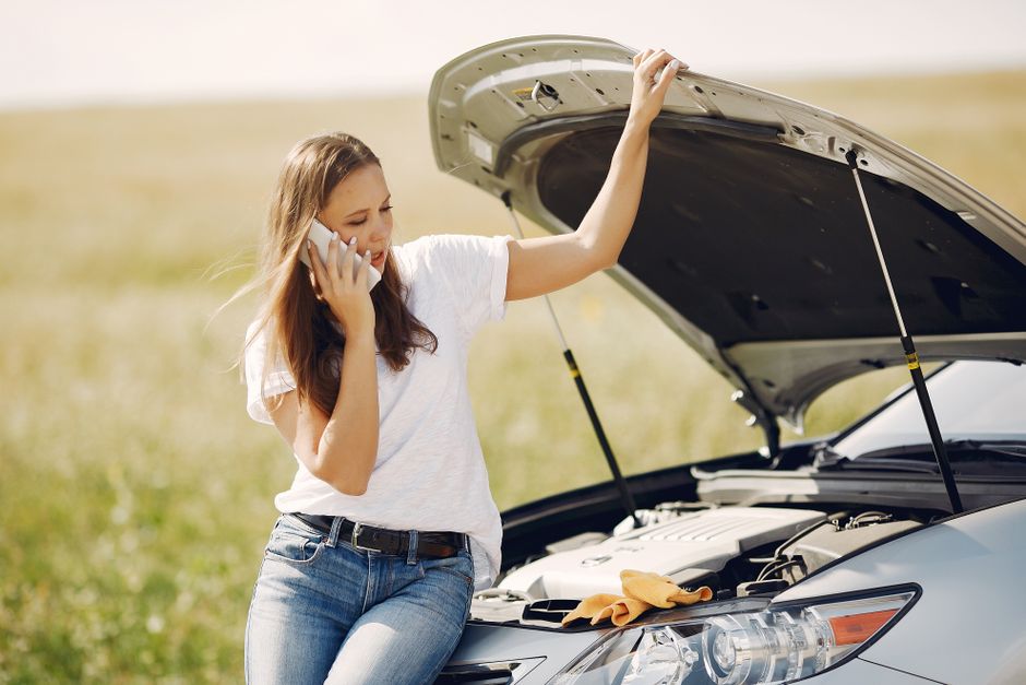 Worried young female driver in white t shirt and jeans speaking on phone while leaning on broken car with open hood asking for help with repair against green field in sunny summer day